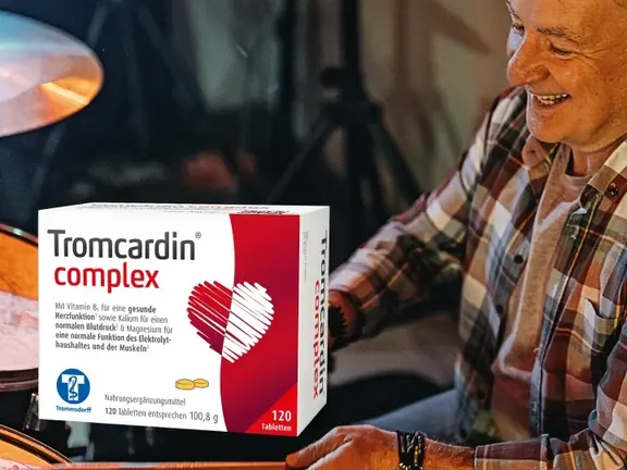 Ein lächelnder älterer Mann, der Schlagzeug spielt, hält eine Schachtel mit Tromcardin complex Nahrungsergänzungsmitteln im Vordergrund.